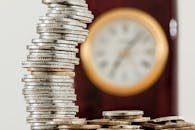 A close-up image of stacked coins with a blurred clock, symbolizing time and money relationship.