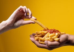A close-up of hands sharing a delicious pile of french fries with ketchup against a yellow background.