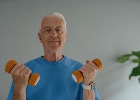 Elderly man in blue shirt lifting dumbbells and smiling indoors, promoting active lifestyle.